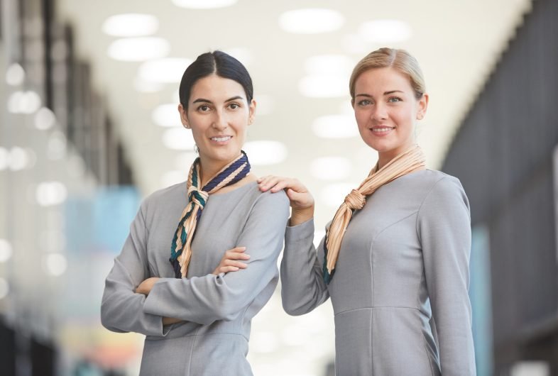 Wast up portrait of two elegant flight attendants looking at camera and smiling while posing in airport