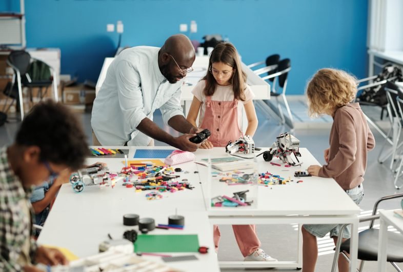 Young teacher bending over table with constructor details and schoolgirl while helping her create new electronic robot at lesson
